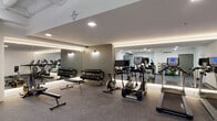 This fitness center is organized into two distinct zones connected by a glass sliding door entrance. The main workout area features a polished, modern aesthetic with a smooth white ceiling, recessed and cove lighting that runs along the perimeter, and large wall-to-wall mirrors that visually expand the space. Durable rubber flooring covers the main gym floor, providing a practical surface for a comprehensive selection of equipment. Cardio machines, including treadmills, ellipticals, and a stationary bike, are arranged in a row along the mirrored wall, while a rowing machine and weight benches occupy the open floor. A fully stocked dumbbell rack and a cable weight machine complete the strength training offerings.

An adjacent functional training room, separated by full-height glass panels, offers a more open, industrial-style setting with an exposed ceiling revealing ductwork and linear LED lighting overhead. This secondary space is equipped with stability balls, medicine balls, resistance accessories, and a cable station, making it well-suited for dynamic or group training sessions. The corridor leading to the facility features a patterned carpet, providing a visual transition into the fitness environment.