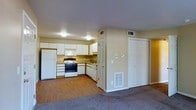 This single-floor space offers a practical, open layout where the kitchen and living area flow seamlessly into one another. The kitchen is equipped with white cabinetry featuring wood-trimmed accents, providing ample upper storage along the walls. Appliances include a white refrigerator, a gas/electric range with an overhead microwave, and a dishwasher, all set against warm yellow-toned walls that add a bright, welcoming feel. A sink is positioned beneath a window, allowing natural light into the cooking area. The kitchen floor transitions from hardwood-style flooring into carpeted living space, clearly delineating the two zones. A wall-mounted thermostat and a floor-level heating vent indicate independent climate control within the unit. The living area features neutral carpet and is bordered by two doors — one appearing to lead to an interior room and another serving as a closet with bifold doors. Overhead flush-mount lighting and HVAC vents in the ceiling ensure year-round comfort. The overall layout is efficient and functional, making good use of the available space across a single level.