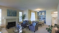 This well-appointed single-floor space showcases a thoughtfully designed open-concept layout that seamlessly connects the living and dining areas. The living area is anchored by a sleek fireplace and features plush upholstered armchairs in soft blue-grey tones, complemented by a reflective glass coffee table that adds a touch of sophistication. Multiple windows dressed with elegant gold curtains and white blinds flood the space with generous natural light, creating a bright and welcoming ambiance. Lush indoor plants add a refreshing, organic element to the décor. The dining area transitions naturally from the living space and is highlighted by a bold blue accent wall adorned with framed artwork and a set of open built-in shelves displaying curated décor pieces. A pendant light fixture overhead provides warm, focused illumination for the dining table and chairs. The overall color palette — a harmonious blend of soft creams, warm golds, and cool blues — lends the space a balanced and inviting character. The neutral carpeted flooring and smooth ceilings tie the rooms together, making the space feel cohesive, comfortable, and ideal for both everyday living and entertaining.