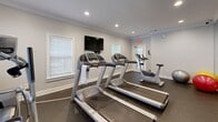 Located at 1000 Arbor Keats Dr in Simpsonville, SC, this fitness center offers a well-appointed exercise space designed to support a well-rounded workout routine. The room features durable dark rubber flooring well-suited for high-impact activity, and is finished with elegant crown molding and neutral gray walls that lend a clean, polished aesthetic.

The space is equipped with multiple treadmills and an elliptical machine for cardiovascular training, along with an upright stationary bike for lower-impact options. Stability balls are also available for core and flexibility exercises. A wall-mounted flat-screen television is positioned for easy viewing during workouts. A large floor-to-ceiling mirror spans one wall, visually expanding the room and allowing users to monitor their form.

Natural light streams in through several windows fitted with white blinds, creating a bright and energizing atmosphere. Recessed ceiling lighting provides consistent illumination throughout. A glass-paned door offers direct access to the outdoors, adding ventilation and a connection to the surrounding greenery. The overall design is functional yet refined, making this fitness center a convenient and comfortable amenity for residents of the Arbor Keats community.