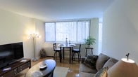 This single-floor space offers a welcoming and functional layout that seamlessly blends living and dining areas into one cohesive environment. The combined living and dining area is a standout feature, anchored by a large picture window dressed with vertical blinds that floods the room with natural light and provides views of the surrounding outdoor scenery. The light-toned tile flooring adds a clean, polished look throughout the space, reflecting the abundant daylight and contributing to an airy, open ambiance. The dining area accommodates a compact round table with bar-height seating, making efficient use of the space near the window. The living area is comfortably appointed with a cushioned sofa and a oval coffee table, creating a relaxed and inviting setting. A stylish multi-arm floor lamp adds a decorative touch while providing additional ambient lighting. Neutral wall tones throughout keep the space feeling bright and versatile. A potted plant near the window introduces a natural element, enhancing the overall freshness of the interior. The thoughtful layout and well-placed furnishings make this a practical and comfortable space suited for everyday living.