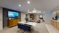 This thoughtfully designed amenity floor offers residents a sophisticated and multifunctional environment suited for both relaxation and productivity. The central lounge area serves as the heart of the space, featuring a striking marble-top dining table surrounded by navy blue velvet chairs with gold-tone legs, a large wall-mounted television, and a warm wood credenza. Recessed ceiling lighting and a sculpted tray ceiling detail with a pendant fixture add architectural depth and a refined ambiance throughout.

Adjacent to the lounge, a library nook provides a curated reading and lounging area complete with a full floor-to-ceiling wooden bookcase, leather accent chairs, and an illuminated tray ceiling. This area transitions seamlessly into a private workspace with a dedicated desk and teal accent wall, ideal for focused work.

A mezzanine-level seating area overlooks the floor below through elegant metal railings, furnished with white rounded lounge chairs and low circular coffee tables beneath statement circular pendant lights, offering a bright and airy retreat with natural light from nearby windows.

The corridor features built-in cabinetry with warm wood finishes and illuminated niches housing business amenities including a multifunction printer and workstation. A quiet lounge with a neutral upholstered sofa and curated gallery wall rounds out the space, connected to a private conference or study room with a built-in desk. Soft carpeting and large-format tile flooring tie the entire floor together cohesively.
