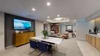This thoughtfully designed amenity floor offers residents a sophisticated and multifunctional environment suited for both relaxation and productivity. The central lounge area serves as the heart of the space, featuring a striking marble-top dining table surrounded by navy blue velvet chairs with gold-tone legs, a large wall-mounted television, and a warm wood credenza. Recessed ceiling lighting and a sculpted tray ceiling detail with a pendant fixture add architectural depth and a refined ambiance throughout.

Adjacent to the lounge, a library nook provides a curated reading and lounging area complete with a full floor-to-ceiling wooden bookcase, leather accent chairs, and an illuminated tray ceiling. This area transitions seamlessly into a private workspace with a dedicated desk and teal accent wall, ideal for focused work.

A mezzanine-level seating area overlooks the floor below through elegant metal railings, furnished with white rounded lounge chairs and low circular coffee tables beneath statement circular pendant lights, offering a bright and airy retreat with natural light from nearby windows.

The corridor features built-in cabinetry with warm wood finishes and illuminated niches housing business amenities including a multifunction printer and workstation. A quiet lounge with a neutral upholstered sofa and curated gallery wall rounds out the space, connected to a private conference or study room with a built-in desk. Soft carpeting and large-format tile flooring tie the entire floor together cohesively.