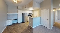 This single-floor unit is finished in a consistent neutral gray-and-beige palette that lends a cohesive, understated aesthetic throughout. The living area is generously proportioned, featuring plush wall-to-wall carpeting and vaulted ceilings that add an airy quality to the space. A ceiling fan provides comfort, while a private exterior door opens to an outdoor area, bringing in natural light through flanking windows fitted with horizontal blinds.

The kitchen is open to the main living space and is anchored by a peninsula counter that provides both workspace and a casual breakfast bar. White cabinetry extends to the ceiling for ample storage, and stainless steel appliances—including a refrigerator, microwave, and range—are complemented by light countertops and wood-look flooring. A double stainless steel sink adds functionality to the layout.

Multiple bedrooms share a similar design language: neutral carpet, blinds-covered windows that bring in natural light, and clean white baseboards. The full bathroom is well-appointed with a white vanity, backlit mirror, wood-look tile flooring, and a tub-shower combination finished in white subway tile. Towel bars and a linen area complete the functional layout of this bathroom.