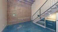Located at 631 Campbell Ave SW in Roanoke, Virginia, this two-level loft showcases a compelling fusion of industrial character and modern design. The main floor makes a strong first impression with rich, polished blue-toned concrete floors that reflect light beautifully, and a prominent exposed brick accent wall that speaks to the building's historic roots. Soaring ceilings amplify the sense of openness, while a contemporary staircase with light wood treads and a steel grid railing leads to the upper level. The kitchen is thoughtfully appointed with sleek two-tone cabinetry, stainless steel appliances including a refrigerator, microwave, and dishwasher, complemented by clean countertops and a functional layout. The full bathroom features a tub-shower combination, a dark wood vanity with ample storage, and a mirror, all set atop the same signature concrete flooring. Ascending to the mezzanine level, the design shifts to warm wide-plank hardwood flooring beneath a stunning exposed wood beam ceiling with skylights that bathe the space in natural light. A set of modern sliding panel doors with a sleek black frame adds a refined architectural detail to the upper level, which overlooks the main floor through the open steel railing. This loft offers a distinctive urban living experience rich in character and thoughtful detail.