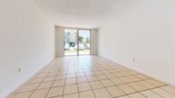 Located at 8920 NW Eighth St in Miami, FL, this 2-bedroom, 2-bathroom unit offers a functional single-floor layout with distinct flooring choices that define each area. The open living room is finished with large neutral ceramic tiles and features a sliding glass door that fills the space with natural light and provides direct access to the outdoors, offering views of the surrounding landscaped grounds. A central hallway connects the main living areas to the private quarters. The kitchen is positioned adjacent to the living room and is equipped with a refrigerator and overhead lighting. Both bedrooms feature light wood-style flooring and large windows that invite natural light while framing outdoor views of the community grounds. The generously sized primary bedroom provides ample space for furnishings. The two bathrooms are finished with tile flooring; one includes a full soaking tub with a tiled surround accented by a decorative mosaic border, paired with a vanity featuring cabinetry and a countertop. The textured ceilings are consistent throughout the common areas, and central air conditioning is present. The overall palette of neutral whites and warm tones gives the unit a clean, bright ambiance throughout.