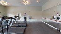 This fitness center offers a clean, functional workout environment designed to support a variety of exercise routines. The space is finished with durable dark rubberized flooring, providing a safe and practical surface for cardio and strength training activities. A ceiling fan with integrated lighting keeps the area comfortable and well-lit during workouts.

The room is equipped with Precor treadmills positioned toward the front, offering users high-quality cardio equipment for their training sessions. A dumbbell rack lines the back wall, stocked with a range of free weights to accommodate different fitness levels. A full-length mirrored wall on one side of the room enhances the sense of space while allowing users to monitor their form during exercise. A wall-mounted ballet barre runs along the mirrored section, making the area suitable for stretching, balance training, or barre-style workouts.

The walls are painted in a neutral light tone, contributing to a calm and focused atmosphere. A doorway leads to an adjacent area, suggesting additional amenities nearby. Informational postings are displayed on the wall, keeping users informed of any facility guidelines. Overall, the space is thoughtfully arranged to maximize usability while maintaining an open, airy feel.