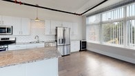 This single-floor loft-style residence offers a striking blend of industrial character and contemporary design. Exposed black pipe systems and large silver ductwork run overhead throughout the open living areas, lending an authentic urban aesthetic against crisp white walls and high ceilings. The expansive main living area features wide-plank hardwood flooring and a modern shiplap-surround fireplace, creating a warm yet minimalist focal point. A dramatic curved bay of floor-to-ceiling windows floods the space with natural light and offers open views of the surrounding streetscape.

The gourmet kitchen is outfitted with sleek white cabinetry, a classic white subway tile backsplash, granite countertops, and a large central island. Stainless steel appliances—including a gas range, built-in microwave, and French door refrigerator—are complemented by a wine cooler, making the kitchen as functional as it is stylish. Pendant lighting adds warmth to the space.

The bedrooms feature plush carpet flooring and generous natural light. The primary bathroom includes a double-sink granite vanity, a glass-enclosed shower, a heated towel rack, and warm tile flooring. A secondary bathroom offers a stacked in-unit washer and dryer alongside a full bath, adding practical convenience to this well-appointed space.