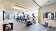 Located at 3040 Oasis Grand Blvd in Fort Myers, Florida, this professionally appointed fitness center offers a comprehensive workout environment designed to meet a wide range of training needs. The space is defined by its expansive floor-to-ceiling window walls that flood the interior with natural light while framing scenic outdoor views of the surrounding Fort Myers landscape, including palm trees and open skies.

The cardio zone is generously equipped with a lineup of SportsArt treadmills, Cybex elliptical trainers, and upright cycling machines, all positioned along the windowed walls to allow exercisers to enjoy the views during their workouts. Mounted flat-screen televisions provide additional entertainment options throughout this area.

The strength training section is equally well-appointed, featuring a robust selection of Cybex selectorized machines, a dedicated free weights area with a full rack of dumbbells, barbells with weight plates, kettlebells, and stability balls. Large wall-to-wall mirrors amplify the sense of space and allow for proper form monitoring during exercises.

The facility's interior design is clean and modern, with a neutral color palette, recessed and linear ceiling lighting, warm wood-toned accent panels, and framed artwork adorning the walls. The overall ambiance is bright, open, and motivating — a premium amenity space built for serious fitness pursuits.