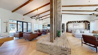 Located at 24720 Morton Ranch Rd in Katy, TX, this expansive single-floor living space delivers an impressive blend of rustic charm and modern sophistication. The open-concept layout is anchored by a dramatic double-sided brick fireplace and column structure that serves as the architectural centerpiece of the room, dividing the space into distinct yet connected zones.

Soaring vaulted ceilings with exposed wood beams span the entire area, creating a grand sense of height and openness. The floors throughout are finished in light-toned herringbone hardwood, adding warmth and visual texture. Large black-framed windows and sliding glass panel doors flood the interior with abundant natural light while offering views of the surrounding exterior.

The space is thoughtfully organized into several functional areas. A lounge zone features rich leather seating arranged around the fireplace, while a separate seating nook with built-in shelving and arched architectural detailing provides a quieter retreat. A wet bar area with tiled accents, open shelving, and bar seating serves as a social hub, complemented by a billiards table beneath a statement pendant chandelier. A dining area with a round table sits against a teal shiplap accent wall adorned with curated artwork. A glass-partitioned room visible through sliding doors adds a versatile, private gathering space to the overall layout.