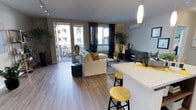 This contemporary single-floor residence showcases a thoughtfully designed open-concept layout that seamlessly connects the kitchen, dining, and living areas into one cohesive, flowing space. The kitchen features a striking granite or stone-composite countertop island with gold-toned bar stools, complemented by a sleek pendant light overhead — a stylish focal point that anchors the entertaining space. Rich wood-look flooring runs throughout, lending warmth and continuity to the interior.

The living area is generously proportioned, bathed in natural light streaming through large floor-to-ceiling windows dressed with dramatic dark curtains. A sliding glass door leads directly to a private balcony, offering outdoor views and enhancing the sense of openness. The space is further elevated by a mini-split climate control unit and a recessed track lighting system, ensuring both comfort and ambiance.

The living room is styled around a plush cream sectional sofa accented with mustard throw pillows, centered on a woven area rug. Lush indoor plants and curated décor add a welcoming, organic touch throughout. A dedicated media area with a dark console completes the space. Overall, the residence exudes a modern, airy aesthetic with a warm and inviting character.