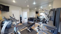 This fitness center offers a thorough and well-organized workout environment designed to support a full range of exercise routines. The space features light wood-style flooring throughout, with protective rubber matting strategically placed beneath equipment areas. Recessed lighting illuminates the room evenly, while multiple windows along the perimeter allow natural light to filter in, creating a bright and energizing atmosphere.

A large wall of floor-to-ceiling mirrors expands the sense of space and allows for proper form checks during exercise. The strength training area is comprehensively stocked, including a full rack of dumbbells spanning a wide weight range, a multi-function cable and weight machine by Paramount, and an adjustable workout bench. Cardio equipment includes an Octane elliptical trainer, a treadmill, a True stationary bike, and an additional elliptical machine.

A wall-mounted flat-screen television provides entertainment during workouts, alongside colorful artwork that adds a touch of personality to the space. A standing water cooler is conveniently positioned near the entrance for easy hydration access. The fitness center connects to an adjacent entryway with glass-paneled doors, offering a welcoming transition into the facility. Informational signage is posted for user guidance, making this a practical and user-friendly workout space.