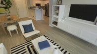 This thoughtfully designed single-floor space offers a clean, modern aesthetic anchored by an open-concept floor plan that effortlessly connects the living area and kitchen. Light-toned hardwood-style flooring runs throughout, lending a sense of continuity and warmth to the interior.

The living area is centered around a striking built-in entertainment unit finished in crisp white, featuring open shelving for display and closed cabinetry below — a combination of style and practicality. The space accommodates comfortable seating and is accented by a bold black-and-white striped area rug that adds a graphic, contemporary touch.

The kitchen is positioned adjacent to the living area, separated by a breakfast bar that doubles as a casual dining and gathering spot. The kitchen is outfitted with stainless steel appliances, including a refrigerator and microwave, set against white cabinetry with a dark countertop that provides an elegant contrast. The open sightlines between the kitchen and living space create a cohesive, sociable environment ideal for both everyday living and entertaining.

The overall ambiance is light, airy, and modern, with a neutral color palette of whites and grays punctuated by navy blue accents. The space feels well-organized and inviting, reflecting a clean, contemporary design sensibility throughout.