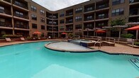 Located at 4000 City Walk Way in Charlottesville, Virginia, this amenity space offers a well-appointed environment designed for comfort and community. The outdoor area is anchored by a generously sized resort-style swimming pool with a connected spa, both surrounded by a paved deck furnished with seating and shaded umbrella stations. The pool is framed by the multi-story building on multiple sides, creating a welcoming courtyard atmosphere.

Inside, the lounge area showcases thoughtful interior design with a cohesive warm palette. Wood-look tile flooring runs throughout, complemented by rich dark wood trim and architectural accents including vertical slatted wall panels. A standout feature is the built-in fireplace nook, finished in vibrant yellow mosaic tile with a chunky wood mantel and a padded bench seat — a cozy focal point within the space.

The seating area is elegantly appointed with an upholstered banquette featuring a textured fabric backdrop, decorative pillows, and stylish accent furniture including ottomans and a metal side table. White ceramic floral wall art adds a decorative touch overhead. The space also includes a corridor with bar-height seating along a counter, recessed and pendant lighting throughout, and glass-paneled entry doors — all contributing to a polished, social atmosphere ideal for residents and guests.