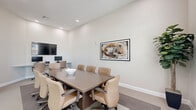 Located at 5353 N US Highway 27 in Ocala, Florida, this professional office space is thoughtfully designed to support productive meetings and collaborative work. The room centers around a substantial rectangular conference table finished in warm wood tones, surrounded by cushioned tan leather rolling chairs that comfortably seat a full team. Recessed lighting provides consistent, even illumination throughout, while oversized windows along one wall invite generous natural light and outdoor views, enhancing the room's open and airy feel. The elevated ceiling height adds a sense of spaciousness that belies the room's compact footprint. Neutral, light-toned walls create a clean and focused atmosphere, accented by a striking framed abstract artwork that adds a touch of visual interest. A wall-mounted flat-screen display and an additional monitor station along one wall ensure the space is fully equipped for presentations and video conferencing. Glass partition walls with sliding panels connect the office to adjacent areas, maintaining visual openness while offering the option for privacy. A decorative fiddle-leaf fig plant introduces a natural element, softening the professional aesthetic. The carpeted floor, laid over a subtle area rug beneath the conference table, contributes to a refined and comfortable environment suited for professional gatherings.