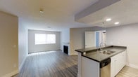 This single-floor, 2-bedroom, 2-bathroom residence offers a well-designed open-concept layout that seamlessly connects the kitchen and living areas. The living room is a welcoming space anchored by a classic fireplace and a large window that allows natural light to fill the room, highlighting the wood-style plank flooring that runs throughout. The neutral gray-toned walls and white trim contribute to a clean, contemporary aesthetic.

The kitchen is positioned at the front of the open layout and features a breakfast bar peninsula with dark countertops, providing both additional prep space and a casual seating area. A stainless steel sink and integrated appliances complete the functional kitchen setup. The peninsula design ensures the kitchen remains visually connected to the living space, enhancing the overall sense of openness.

A paneled door visible from the main living area leads to the private quarters, where two bedrooms and two full bathrooms are accommodated. The single-floor plan offers convenient, step-free living throughout. Overall, the space combines practical functionality with a modern, understated design suitable for comfortable everyday living.