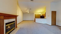 Located on S Moorland Rd in New Berlin, Wisconsin, this single-floor unit showcases an open and airy layout defined by soaring vaulted ceilings in the main living area. The spacious living room serves as the central gathering space, anchored by a classic wood-manteled fireplace with a glass-front insert, adding warmth and a traditional focal point to the room. Plush wall-to-wall carpeting runs throughout the living and bedroom areas, contributing to a comfortable, quiet atmosphere.

The kitchen is thoughtfully designed with an open-concept arrangement that connects seamlessly to the living area via a curved breakfast bar. Warm oak cabinetry lines the walls, complemented by light countertops and black appliances, including an electric range and dishwasher. A chandelier overhead adds a refined touch to the dining space adjacent to the kitchen.

The bedrooms are well-proportioned, carpeted spaces with ample wall area and closet access. Natural light filters in through windows dressed with blinds. The full bathroom features oak vanity cabinetry, a tub-shower combination, tile flooring, and generous counter space. A wood baluster staircase detail is also visible, adding a classic architectural element to the overall design.