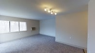 This single-floor space presents a straightforward, functional layout finished in a neutral color palette that creates a calm and adaptable atmosphere. The main living area is carpeted in a medium-toned gray, providing a soft, comfortable underfoot feel throughout the room. Walls are painted in a clean, light gray-white tone, contributing to a bright and open ambiance.

Overhead, a contemporary multi-head track lighting fixture with warm-toned bulbs serves as the room's primary light source, casting even illumination across the space. A window fitted with horizontal blinds allows for natural light while maintaining privacy, and a wall-mounted window air conditioning unit provides climate control directly within the room.

White baseboards run cleanly along the perimeter, adding a crisp contrast to the carpeted floors. The open layout of the room allows for flexible furniture arrangement, making it suitable for a variety of uses. Overall, the space offers a clean, move-in-ready environment with practical features and a neutral aesthetic that can accommodate a range of personal styles and furnishing choices.