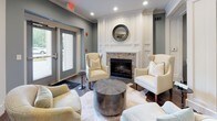 Located at 3112 Maybank Hwy on Johns Island, South Carolina, this refined single-floor clubhouse offers a seamlessly connected layout designed for both relaxation and social gatherings. Rich, dark hardwood flooring runs throughout the entire space, anchoring a cohesive and sophisticated aesthetic defined by soft blue-gray walls, crisp white millwork, and decorative columns.

The main lounge area is generously proportioned, featuring multiple seating arrangements centered around a large area rug. Twin flat-screen televisions are mounted on a shiplap accent wall, while recessed lighting and crown molding add a polished finish overhead. French doors provide access to adjacent areas, reinforcing the open, flowing floor plan.

A dedicated fireplace lounge offers an intimate retreat, anchored by a stone-surround fireplace framed with white shiplap paneling and a circular mirror above the mantel. Cream wingback chairs and layered seating create a warm, inviting atmosphere in this corner of the space.

The kitchenette is thoughtfully appointed with white shaker cabinetry, a striking chevron-patterned tile backsplash, gray quartz countertops, stainless steel appliances including a wine cooler and microwave, and a spacious bar-height counter with seating. A separate high-top dining nook with a wall-mounted television provides an additional casual gathering option. Together, these areas combine comfort, functionality, and refined design.