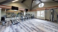 This fitness center is housed in a single-story space distinguished by striking vaulted ceilings with exposed dark wood beams that soar overhead, lending an open, airy atmosphere. A large decorative round window near the ceiling peak allows natural light to flood the interior, complemented by additional standard windows offering outdoor views, including a glimpse of a pool area accessible through glass-panel French doors.

The interior is finished with durable patterned flooring throughout, accented by terracotta-toned horizontal stripes along the upper walls that add a bold design element. Full-length mirrors line one wall, visually expanding the space and supporting proper form during workouts.

The cardio zone features multiple treadmills and elliptical trainers, each equipped with individual monitors. Wall-mounted flat-screen televisions are positioned for easy viewing during exercise. The strength training area includes a multi-function cable machine, a rack of dumbbells with a weight bench, and a dedicated functional fitness station stocked with medicine balls, resistance bands, stability balls, and yoga mats. The open floor plan allows for flexible use of the space, making it suitable for both structured workouts and group exercise activities.
