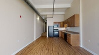 Located at 22 S Saint Clair St in downtown Dayton, Ohio, this single-floor unit showcases a distinctive industrial-loft aesthetic with soaring ceilings that draw the eye upward to exposed ductwork and structural beams running overhead. The open layout creates a generous sense of space, with warm wood-tone laminate flooring extending throughout the main living areas, grounding the industrial character with a welcoming feel.

The kitchen is well-appointed, featuring abundant warm-toned wood cabinetry with modern hardware, granite-style countertops, a double stainless steel sink, and a full suite of black appliances including a refrigerator, electric range with microwave above, and dishwasher. Upper and lower cabinets provide ample storage, and under-cabinet lighting adds functional illumination to the workspace.

A separate bedroom area is finished with plush carpet and features a clerestory window that allows natural light to filter in while maintaining privacy. Exposed ductwork continues through this space, reinforcing the loft-style character throughout.

The bathroom is clean and functional, offering a dark-finished vanity with a framed mirror, a single-basin sink, and a bathtub/shower combination, all set on wood-look flooring. The overall ambiance balances urban industrial design with practical everyday comfort.