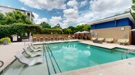 Located at 2841 Manorcrest Ct in Raleigh, NC, this outdoor amenity space is designed to deliver a resort-style experience for residents. At its centerpiece is a generously sized swimming pool with crystal-clear turquoise water, accessible via stainless steel entry rails and flanked by wide concrete pool decking. A well-appointed sun deck lines one side of the pool, outfitted with a row of sleek chaise lounges accented with bright yellow cushions, providing ample space for relaxation and sunbathing. Shade options include market umbrellas and a covered outdoor lounge area featuring a ceiling fan, exposed brick columns, and comfortable seating arranged on a bold patterned area rug — ideal for socializing in cooler comfort. Lush landscaping, including tall columnar trees, flowering shrubs, and potted greenery, frames the space with vibrant color and privacy. An outdoor grilling and dining area with additional seating is positioned nearby, enhancing the space's entertaining potential. The pool courtyard is enclosed by the building's multi-story brick and board-and-batten facade, with resident balconies overlooking the area. Natural light floods the space throughout the day, creating a bright and welcoming atmosphere that seamlessly blends indoor and outdoor living.