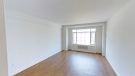 This single-floor space presents a clean, minimalist aesthetic defined by crisp white walls and smooth hardwood flooring that lends warmth and continuity throughout the interior. The layout is open and uncluttered, allowing for flexible use and easy configuration to suit a variety of needs.

A prominent multi-pane window serves as a central architectural feature, drawing in generous amounts of natural light that brightens the entire space. The window frames a view of the surrounding urban environment, offering a glimpse of neighboring multi-story buildings that speak to the city setting. Below the window, a built-in ventilation unit is neatly integrated into the wall, maintaining the space's tidy appearance.

The ceiling is smooth and uniformly white, with subtle structural detailing visible in the form of a raised soffit near the window wall, adding a layer of architectural character without disrupting the overall sense of openness. Crown molding and clean baseboards run throughout, contributing to the polished, well-finished feel of the space.

Overall, the space conveys a sense of calm and simplicity, making it a versatile and inviting environment suited for residential or professional use.