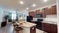 This single-floor residence offers a thoughtfully designed open-concept layout that seamlessly connects the kitchen, dining, and living areas. The kitchen is a standout feature, equipped with rich dark wood cabinetry, light stone-toned countertops, a full suite of black appliances including a side-by-side refrigerator with ice and water dispenser, an electric range, over-the-range microwave, and a double sink. A curved breakfast bar provides additional seating and serves as a natural divider between the kitchen and the adjoining living space. The living area is bright and inviting, featuring light wood-style flooring, a ceiling fan, and large windows dressed with teal curtains that allow natural light to fill the room. The bedroom is carpeted for comfort, includes a ceiling fan, ample natural light from a window with blinds, and is well-proportioned for a full furniture arrangement. The bathroom features matching dark wood cabinetry, a dual-sink vanity with stone countertops, wood-style flooring, and leads directly to a walk-in closet. A dedicated laundry area with a full-size washer and dryer completes the functional layout of this well-rounded residence.