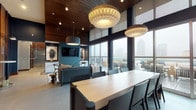 Located at 101–165 E Louisiana St in downtown Indianapolis, this single-floor amenity lounge delivers a refined, contemporary atmosphere defined by thoughtful design and high-end finishes. Soaring ceilings clad in warm wood paneling contrast elegantly with deep navy and teal accent walls, establishing a bold yet inviting palette throughout the space.

Floor-to-ceiling windows and glass sliding doors flood the interior with natural light while framing sweeping views of the Indianapolis skyline. These openings connect seamlessly to an outdoor terrace furnished with seating, extending the usable space beyond the interior.

A generously sized conference-style dining table anchors the central area, illuminated by sculptural pendant lights with distinctive layered shades. Adjacent lounge seating—including a substantial sectional sofa and sculptural accent chairs—creates a comfortable gathering zone centered around a wall-mounted television and curated artwork.

A wet bar and service counter featuring white cabinetry is set against a dramatic Indianapolis skyline mural, offering a functional yet visually striking backdrop. Along one wall, sleek high-top tables with articulating sconce lighting provide additional workspaces or casual seating areas. Large-format stone tile flooring unifies the space, lending a polished, cohesive finish throughout.