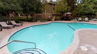 This inviting outdoor community space is designed for relaxation and recreation, centered around a generously sized freeform swimming pool with a classic tiled border and clear blue water. Surrounding the pool, a spacious stamped concrete deck provides ample room for lounge chairs, umbrella tables, and seating arrangements, making it ideal for sunbathing or socializing. Adjacent to the main pool, a dedicated spa offers a serene spot to unwind, nestled among lush greenery and decorative boulders for a natural, resort-inspired feel. A separate BBQ and picnic area is conveniently equipped with a gas grill, picnic table, and waste receptacle, perfect for outdoor gatherings and casual dining. Mature trees and well-maintained landscaping with shrubs, ground cover, and garden beds are thoughtfully distributed throughout the space, providing shade, privacy, and a tranquil atmosphere. Concrete pathways connect each amenity zone, allowing easy navigation across the grounds. The surrounding multi-story residential buildings, finished in warm tan siding, frame the courtyard and contribute to the enclosed, community-oriented feel of the space. Overall, this outdoor area delivers a comfortable and functional environment with a strong emphasis on leisure and community living.