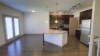 This two-floor space showcases a thoughtfully designed open-concept main level where the kitchen and living area flow seamlessly together. The kitchen is appointed with rich, dark espresso cabinetry that provides a striking contrast against the light granite countertops. A central island with an integrated sink serves as both a functional workspace and a natural gathering point, finished with a granite surface that mirrors the surrounding counters. Stainless steel appliances, including a range and built-in microwave, add a contemporary touch, while a decorative mosaic tile backsplash introduces subtle color and texture to the space. Lighting is layered effectively, combining pendant lights suspended over the island, a track lighting system, and a ceiling-mounted fixture in the living area to ensure the space is well-lit throughout. The hardwood-style flooring runs continuously across the open area, grounding the design with warmth and cohesion. The living area offers generous open space, providing flexibility for various furniture arrangements. Neutral wall tones throughout keep the palette clean and versatile, contributing to the overall modern and welcoming ambiance of the space.
