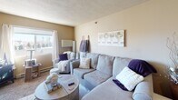 Located at 3120 W Rambler Pl in Sioux Falls, SD, this single-floor unit offers a practical and comfortable layout with a warm, neutral color palette throughout. The open-concept living area is bright and inviting, featuring carpeted flooring and a window that allows natural light to fill the space. The living room accommodates a generous sectional sofa and provides a relaxed atmosphere suited for everyday living.

The kitchen is efficiently designed with warm wood cabinetry, light countertops, and black appliances including a range and over-the-range microwave. A breakfast bar with seating connects the kitchen to the main living area, making the space feel cohesive and functional.

The bedroom is a quiet, carpeted retreat featuring a window with blackout curtains, a closet, and ample room for a full-sized bed and furnishings. The textured ceiling and warm wall tones carry throughout the space, creating a consistent aesthetic.

The bathroom is finished with tile flooring, a shower-tub combination with a full curtain, a vanity with cabinet storage, and warm-toned walls. Additional open shelving provides practical storage space.

Overall, this unit presents a cozy, well-maintained living environment with a straightforward floor plan that maximizes functionality in a single-story layout.