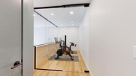 Located at 829 Royal Ave SW in Calgary, this thoughtfully designed single-floor amenity space offers a clean, modern aesthetic across its compact footprint. The layout is divided into two distinct functional areas, each finished to a high standard.

The centerpiece is a glass-enclosed fitness room accessed through full-height glass sliding doors with sleek black framing. Inside, light hardwood flooring provides a warm contrast to the crisp white walls and recessed ceiling lighting. The room currently houses stationary spin bikes on protective floor mats, making it a dedicated exercise zone. A built-in wet bar with light wood cabinetry, a white countertop, and a sink sits adjacent to the glass partition, adding practical convenience for the fitness area.

The second area features the same warm hardwood flooring and is finished with soft sage-green accent walls, providing a calm, welcoming atmosphere. A window allows natural light to filter through. This zone connects to a washroom finished in neutral floor tiles, featuring a wall-mounted sink and a round mirror, maintaining the space's clean, contemporary character throughout.

Overall, the space presents a polished, functional environment well-suited as a shared or private amenity suite.