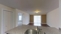 This single-floor residence offers a thoughtfully designed open-concept layout that seamlessly connects the kitchen and living areas. The kitchen is appointed with granite countertops and a double stainless steel sink, providing both functionality and a refined aesthetic. The living area features rich hardwood flooring, which adds warmth and contrast to the neutral cream-toned walls. A bold accent wall in warm tan anchors the living space, lending depth and character to the room. Large sliding glass doors dressed with vertical blinds allow an abundance of natural light to pour in while offering access to the outdoors. A hallway visible from the main living area leads to additional rooms, which feature plush carpeting and benefit from their own natural light sources through well-placed windows. The space is finished with clean white trim and baseboards throughout, contributing to a cohesive and polished look. Overhead lighting is present in the living area, ensuring the space is well-lit at all times of day. The overall ambiance is bright, welcoming, and practical, making it a comfortable and move-in-ready living environment.