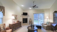 This single-floor home presents a welcoming and well-appointed living space defined by its vaulted ceilings that lend an airy, open feel to the interior. The main living area serves as the heart of the home, anchored by a classic tile-surround fireplace with a clean white mantel — an ideal focal point for both relaxed evenings and entertaining. Above the mantel, the space is designed to accommodate a flat-screen display, blending functionality with traditional aesthetics. A ceiling fan with a light fixture is centrally positioned to provide comfort and illumination throughout the room. Rich, dark wood furnishings complement the neutral-toned walls and light carpeting, creating a warm and inviting ambiance. A large credenza along one wall adds both storage and character, while framed artwork contributes a refined touch. Sliding glass doors on one side of the living area flood the space with natural light and offer direct views of a covered outdoor patio and lush, wooded surroundings — seamlessly connecting the interior to the outdoors. The overall design balances classic architectural details with comfortable, livable finishes, making this a thoughtfully crafted single-level home.