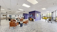 Located at 1707 Owen Dr in Fayetteville, NC, this generously sized single-floor commercial space offers a modern, open-concept layout with a vibrant and welcoming atmosphere. The interior is anchored by distinctive terrazzo flooring that runs throughout, complemented by a drop-panel ceiling with recessed LED lighting that keeps the space bright and airy.

The main open area serves as a central lounge and gathering space, furnished with leather seating clusters, low coffee tables, and open shelving units. Bold purple accent walls add a striking visual identity, while large floor-to-ceiling windows along the perimeter flood the interior with abundant natural light and provide views of the surrounding greenery. A beverage station and wall-mounted television make the lounge both functional and inviting.

A dedicated recreation area features a full-size billiards table and bar-height seating, ideal for casual social interaction. Nearby, a private office is accessed through elegant glass-paned French doors with blue trim, offering a quieter workspace. An eye-catching staircase with dark stone-finish treads, black metal railings, and glass balustrade panels serves as an architectural focal point. Curated artwork is displayed throughout, contributing to the space's creative and professional character. Glass-front entrances on multiple sides enhance accessibility and connectivity to the outdoors.