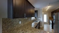 This single-floor space showcases a thoughtfully designed interior with refined finishes and a cohesive aesthetic throughout. The kitchen stands out as a highlight, featuring rich dark espresso cabinetry with sleek brushed nickel hardware that creates a striking contrast against the polished granite countertops. The granite surfaces display a warm blend of earthy tones with dynamic veining, adding both elegance and visual depth to the space. A undermount sink sits beneath a window, allowing natural light to filter into the kitchen workspace, while the matching granite backsplash ties the design together seamlessly. Crown molding along the upper cabinetry and ceiling transitions adds a classic architectural touch, elevating the overall sense of quality and craftsmanship. The light-toned tile flooring provides a bright, neutral foundation that balances the darker cabinetry and contributes to an open, airy feel. A glimpse into the adjoining area reveals louvered closet doors and continued neutral tones, suggesting a consistent design language carried throughout the space. Overall, the interior strikes a balance between contemporary style and timeless detail, creating an environment that is both functional and visually appealing.