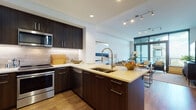 This contemporary single-floor residence offers a thoughtfully designed open-concept layout that seamlessly blends style and functionality. The kitchen is a standout feature, showcasing rich dark-wood cabinetry, light quartz countertops, a large center island with a deep double sink and an elegant industrial-style faucet, and a full suite of stainless steel appliances including a built-in microwave, range, dishwasher, and French door refrigerator. A textured tile backsplash and warm hardwood flooring complete the kitchen's polished aesthetic. The kitchen flows naturally into a combined dining and living area, where track lighting illuminates the space and expansive floor-to-ceiling windows with a glass-panel door flood the room with natural light and provide sweeping city views. The bedroom continues the floor-to-ceiling window theme, offering breathtaking panoramic views of the surrounding cityscape and soft carpeted flooring for comfort. The bathroom is elegantly finished with large-format tile flooring, a dark wood vanity with a white quartz countertop, and a full soaking tub with marble-style tile surround. Directly accessible from the bathroom is an organized walk-in closet, adding a practical and luxurious touch to the private quarters.