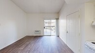 This single-floor unit offers three bedrooms, one bathroom, a kitchen, a hallway, and a closet — all finished with consistent wood-look flooring that runs throughout the space. The main living area is bright and open, featuring vaulted ceilings that lend a sense of added vertical space, a wall-mounted HVAC unit, and a sliding glass door that opens to a private balcony with outdoor views. The textured ceilings and clean white walls create a neutral, move-in-ready aesthetic across the unit. The kitchen is well-equipped with white cabinetry, open shelving, a refrigerator, a range, a dishwasher, a sink, and a flush-panel ceiling light fixture, offering functional workspace along two walls. Each of the three bedrooms maintains the same clean, light-toned palette, with wood-look floors and windows that allow natural light to filter in. The primary bedroom is comfortably sized and includes a wall heating unit. The bathroom is fitted with a tub-shower combination featuring chrome fixtures and a grab bar. A dedicated closet and a connecting hallway complete the thoughtfully organized layout of this compact, efficiently designed unit.