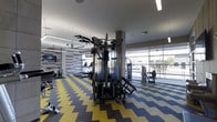 This fitness center is designed to support a comprehensive workout experience within a single, open-plan level. The space is immediately distinguished by its striking yellow and grey chevron-patterned flooring, which adds a dynamic visual energy to the room. At the center of the facility stands a multi-station cable machine, offering a wide range of strength training options for users of varying fitness levels. Surrounding it, a selection of cardio equipment—including treadmills and stationary bikes—lines the perimeter, providing ample options for aerobic exercise. Large floor-to-ceiling windows along one wall flood the space with natural light while offering views of the exterior surroundings, creating an open and airy atmosphere that enhances the workout environment. A textured brick accent wall adds an industrial design element, complementing the modern ceiling with integrated linear lighting and ceiling fans that help maintain comfortable airflow. Mounted screens are visible throughout, likely used for entertainment or fitness programming. The overall layout is spacious and thoughtfully organized, balancing equipment density with ease of movement.