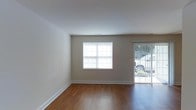 This single-floor space offers a clean, functional layout with a welcoming interior aesthetic. The main living area is defined by rich, dark hardwood flooring that runs throughout, providing warmth and visual continuity. The walls are finished in a soft neutral beige tone, creating a calm and versatile backdrop suitable for a variety of interior styles. A centrally placed window fitted with horizontal blinds allows natural light to filter into the room, brightening the space during daylight hours. Adjacent to the window, a glass door with vertical blinds opens to an outdoor area, offering views of the exterior surroundings and providing an additional source of natural light. The white ceiling is smooth and clean, featuring a ceiling vent that contributes to the room's practical infrastructure. White baseboards frame the perimeter of the room, adding a crisp contrast to the neutral walls and dark flooring. The overall ambiance of the space is modest and move-in ready, with a straightforward design that prioritizes functionality. The open feel of the main room makes it adaptable for various furniture arrangements and uses.