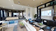 Located in the Carmel Creek neighborhood of San Diego, this single-story clubhouse serves as a central gathering space for its community. The interior showcases a contemporary design aesthetic, with soaring ceilings and recessed lighting that illuminate the open-plan layout throughout. The main lounge area is anchored by a plush cream sofa and woven rattan accent chairs in rich brown and blue tones, arranged around a rustic wood coffee table to create an inviting conversational setting. Potted succulents and lush greenery add natural vibrancy to the space. A striking built-in shelving unit with dark cabinetry and textured stone accenting serves as a focal point along one wall, displaying decorative accessories and offering storage. Light wood-toned flooring runs continuously through the space, complementing the warm neutral wall palette. Toward the rear, an additional lounge or service area is partially visible, defined by geometric decorative screens and exposed beam detailing that add architectural interest. The overall ambiance is polished yet comfortable, balancing modern sophistication with approachable, community-oriented warmth.