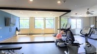 This fitness center offers a bright and functional workout environment designed to support a range of exercise routines. The space is characterized by large windows along one wall, allowing abundant natural light to filter in while providing pleasant views of surrounding greenery. Full-length mirrors are mounted across an adjacent wall, enhancing the sense of openness and making the space feel more expansive. The flooring transitions between a smooth hardwood-style surface in the stretching and open exercise area, and durable rubber matting beneath the cardio equipment, offering both practicality and comfort. True brand elliptical and cardio machines are positioned along the mirrored wall, providing quality equipment for cardiovascular training. Stability and medicine balls are visible in the open floor area, which offers ample room for stretching, balance work, or group exercise. Recessed ceiling lighting supplements the natural light, ensuring the space remains well-lit throughout the day. The overall ambiance is clean, modern, and welcoming, with a neutral color palette of warm beige walls and dark trim contributing to a focused yet comfortable atmosphere well-suited for fitness activities.
