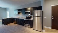 This single-floor unit offers a thoughtfully designed layout that balances functionality with modern aesthetics. The living area is finished with plush wall-to-wall carpeting and neutral gray walls, creating a warm and inviting atmosphere. Multiple windows fitted with horizontal blinds allow natural light to fill the space, enhancing its open and airy feel.

The kitchen is a standout feature, showcasing rich dark cabinetry that extends to the ceiling and is topped with light granite countertops. A full suite of stainless steel appliances includes a refrigerator, electric range, over-the-range microwave, and dishwasher. A built-in diamond-pattern wine rack adds a distinctive design touch, while light hardwood-style flooring grounds the space with warmth.

A dedicated laundry area provides added convenience with a stacked washer and dryer unit, complemented by built-in wire shelving for storage. Adjacent to the laundry, the bathroom features granite-topped dark vanity cabinetry, a full soaking tub with shower, and hardwood-style tile flooring. A walk-in closet with wire shelving is accessible directly from the bathroom, offering generous storage. Throughout the unit, clean white baseboards and consistent finishes create a cohesive, polished look.