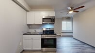 This efficiently designed one-bedroom, one-bathroom unit offers a thoughtful layout across a single floor. The open kitchen and living area form the heart of the space, featuring wood-look laminate flooring in a cool gray tone that extends throughout. The kitchen is equipped with stainless steel appliances, including an electric range, over-the-range microwave, and a single-basin sink, all complemented by white shaker-style cabinetry and dark countertops. The living area benefits from a ceiling fan with integrated lighting, a wall-mounted air conditioning unit, and a baseboard heating system, ensuring year-round comfort. A window with blinds allows natural light to fill the space. The bedroom is a well-proportioned room featuring the same laminate flooring, a window for natural light, and a full-width closet with accordion-style doors providing ample storage. An additional built-in wardrobe cabinet further enhances storage capacity. The bathroom is finished in a clean, neutral palette with white subway tile, a single-sink vanity with a framed mirror, a soaking tub, and a window that brings in natural light. The textured ceiling is consistent throughout, adding a classic touch to the overall design.