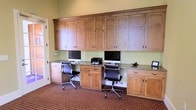 This single-floor business center offers a functional and organized workspace thoughtfully designed for productivity. The room is anchored by an expansive wall of warm-toned, natural wood built-in cabinetry that spans floor to ceiling, featuring arched panel doors with dark hardware and generous upper storage compartments. Below the upper cabinets, a continuous built-in countertop provides ample desk space, accommodating multiple workstations complete with desktop computers, a printer, and additional peripherals. Ergonomic black office chairs are positioned at each station, ensuring comfort during extended use. The lower cabinetry mirrors the upper units, offering additional drawers and closed storage for a clean, clutter-free environment. The space is finished with a patterned carpet in warm, earthy tones that adds a touch of classic character, while soft green walls provide a calm and focused atmosphere. A glass-panel door allows natural light to filter into the room, enhancing the overall brightness. The cohesive design of the built-in millwork and thoughtful layout make this business center a practical and welcoming resource for work or study.