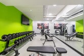 Located at 98 Farley Drive in Guelph, Ontario, this fitness facility offers a thoughtfully designed, single-floor space that accommodates a comprehensive range of workout options. The interior is characterized by a bold and energizing color scheme, with vivid green, blue, and yellow accent walls contrasting against a clean white ceiling and neutral flooring, creating a motivating atmosphere for users.

The free weights area features a full rack of dumbbells arranged along a bright green accent wall, complemented by a wall-mounted flat-screen television and athletic-themed artwork, adding visual interest to the training environment. Adjustable weight benches and a multi-functional cable machine provide ample options for strength training, while a large floor-to-ceiling mirror along one wall enhances the sense of space and allows users to monitor their form.

The cardio zone is lined with a row of treadmills equipped with individual screens, alongside elliptical trainers and stationary bikes. A mounted television on the blue accent wall keeps users entertained during their workouts. Additional equipment, including rowing machines and cable stations, is positioned throughout the space to maximize functionality.

Natural light filters in through windows along one wall, offering views of surrounding greenery and contributing to an open, inviting ambiance. Recessed lighting throughout ensures consistent illumination across the entire facility.