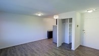 This single-floor studio offers a clean, open layout designed to maximize functionality within a compact footprint. The main living area features smooth hardwood-style flooring in a warm grey-brown tone, complemented by crisp white walls and flush ceiling lighting that keeps the space feeling bright and airy. The open plan seamlessly integrates the living and kitchen areas, where a kitchenette is visible at the rear, equipped with dark cabinetry, a light granite-style countertop, and a full-sized refrigerator — providing essential cooking amenities in an efficient arrangement. A standout feature of this space is the built-in closet, positioned centrally and fitted with a clothing rail and multiple open shelving units above, offering well-organized storage solutions. The door to the closet is a classic bifold panel design, adding a tidy, unobtrusive aesthetic. The overall ambiance is clean, minimal, and move-in ready, with a neutral color palette throughout that lends itself well to personalization. The layout is practical and straightforward, making excellent use of the available floor space while maintaining an uncluttered, open feel.