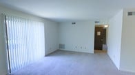 This single-floor residential space offers a straightforward and functional layout designed for comfortable everyday living. The main living area is generously proportioned, featuring wall-to-wall carpet in a neutral tone that adds warmth and softness underfoot. Large vertical blinds line one wall, concealing what appears to be a full-width sliding glass door that allows for an abundance of natural light to fill the room. The walls are finished in a clean, bright white, contributing to an open and airy atmosphere throughout the space. Ceiling-mounted HVAC vents ensure consistent climate control, while a baseboard heating unit along the far wall provides supplemental warmth. Multiple electrical outlets are positioned conveniently along the walls, offering practical utility throughout the living area. A doorway visible at the rear of the main room leads to an additional space, hinting at a connected layout that extends beyond the primary living area. The overall ambiance is clean, minimal, and move-in ready, presenting a blank canvas well-suited for a variety of interior styles and furnishing arrangements.