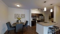 This single-floor residence showcases an open-concept layout that seamlessly connects the kitchen and dining areas. The kitchen is outfitted with rich, dark-toned cabinetry paired with granite countertops, stainless steel appliances — including a French door refrigerator, electric range, and built-in microwave — and a peninsula with an undermount sink. A pendant light hangs above the peninsula, complemented by track lighting overhead, providing ample task illumination throughout the cooking space. The dining area sits adjacent to the kitchen and accommodates a round table with seating for multiple guests. Decorative geometric wall art adds a contemporary touch to the dining space. Crown molding runs along the ceiling perimeter, lending an elegant architectural detail throughout. Dark hardwood-style flooring extends across both areas, offering a warm and cohesive aesthetic. The neutral wall tones and clean white baseboards create a bright, polished atmosphere, while the open floor plan allows natural sightlines across the entire living space, making it feel connected and welcoming.